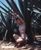 A naked woman kneeling in front of a palm tree.