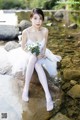 A woman in a white dress sitting on a rock by a river.