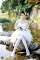 A woman in a white dress sitting on a rock by the water.